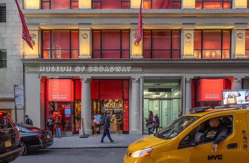 The red-lit entrance of the Museum of Broadway, with a yellow taxi passing on the street outside