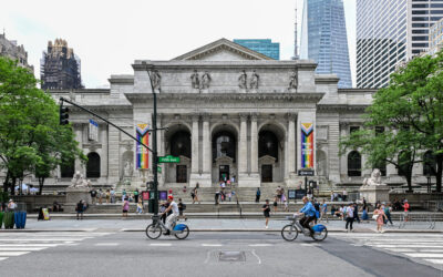 The Fifth Avenue facade of the New York Public Library seen from across the street, Pride banners between the columns and Citi Bike riders passing in front