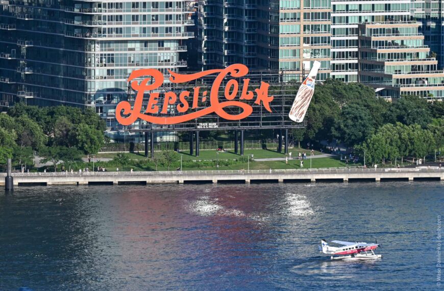 Pepsi-Cola neon sign on Manhattan waterfront with city buildings in background.