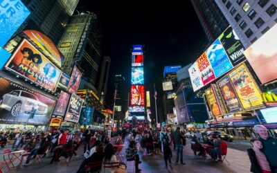 Times Square at night, ringed by illuminated billboards and theatre marquees, with crowds seated on red chairs