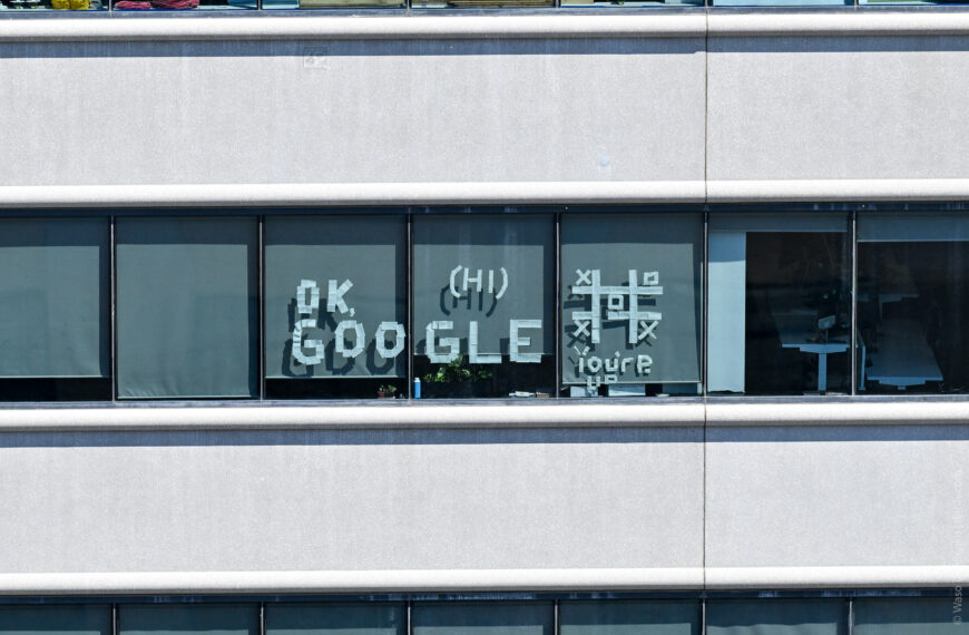 Taped letters in an office window reading OK, GOOGLE (HI) beside a game of tic-tac-toe and the note YOU'RE UP